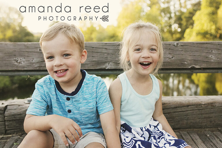 Two children smiling on a wooden bench outside.