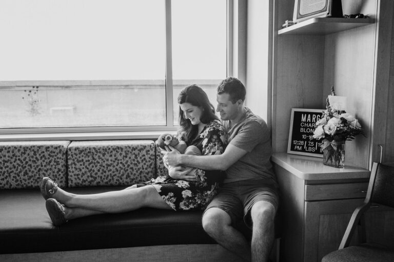 Parents holding newborn by window in hospital room.