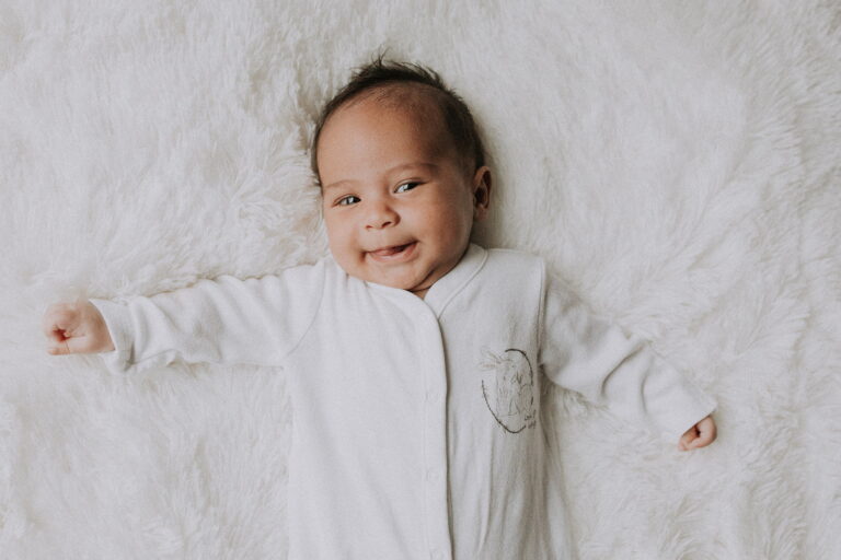 Smiling baby in white outfit on fluffy blanket.