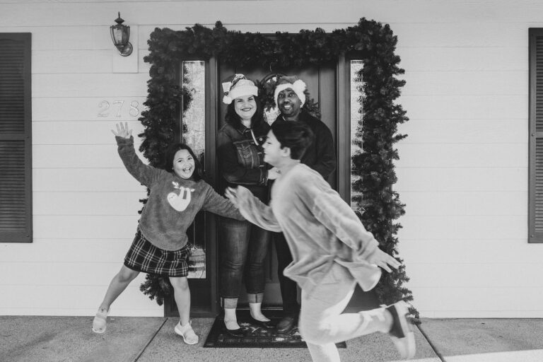 A mom and dad in Santa hats, stand in tranquil composer to contrast with the blur of playing children in this holiday portrait.