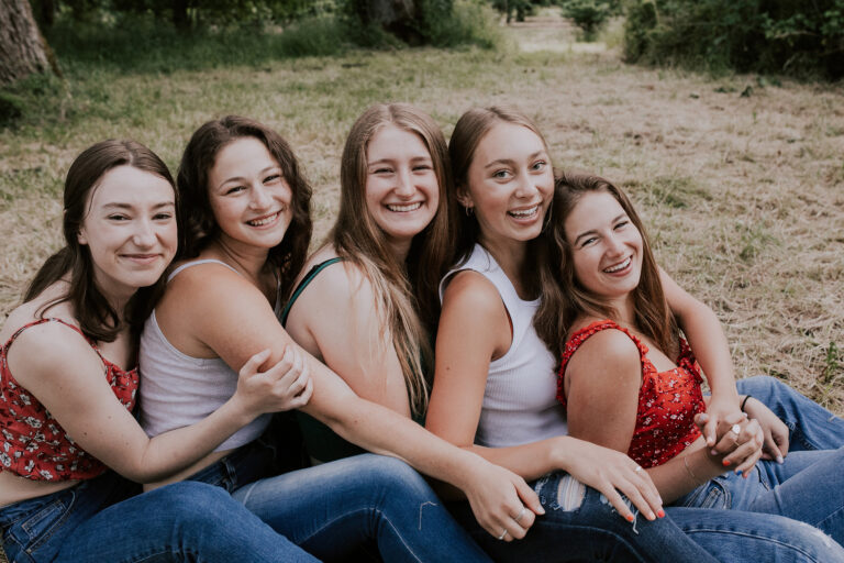 Five friends sitting on grass, smiling outdoors.