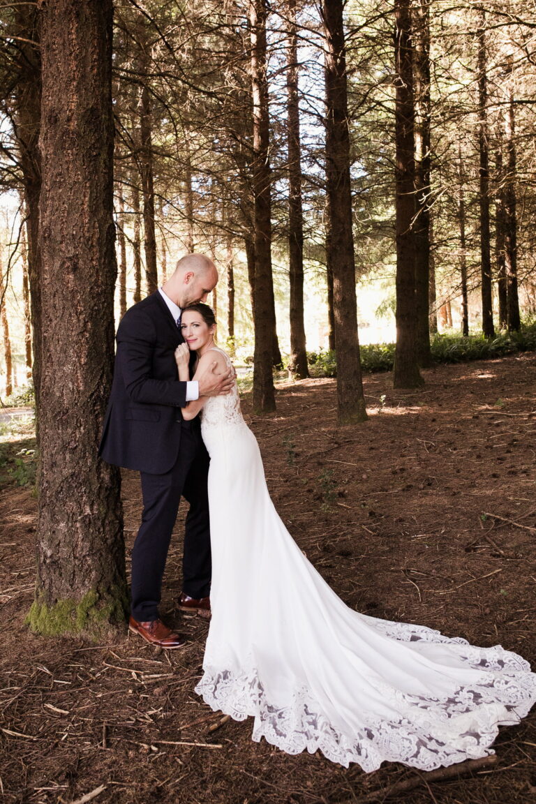 The long white train of a wedding gown takes center-stage as a bride leans into the embrace of her groom in a shaded forest.