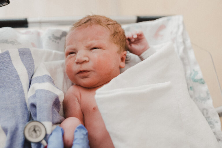A newborn squints into the light of a hospital room as a parent's hand reaches to great them.