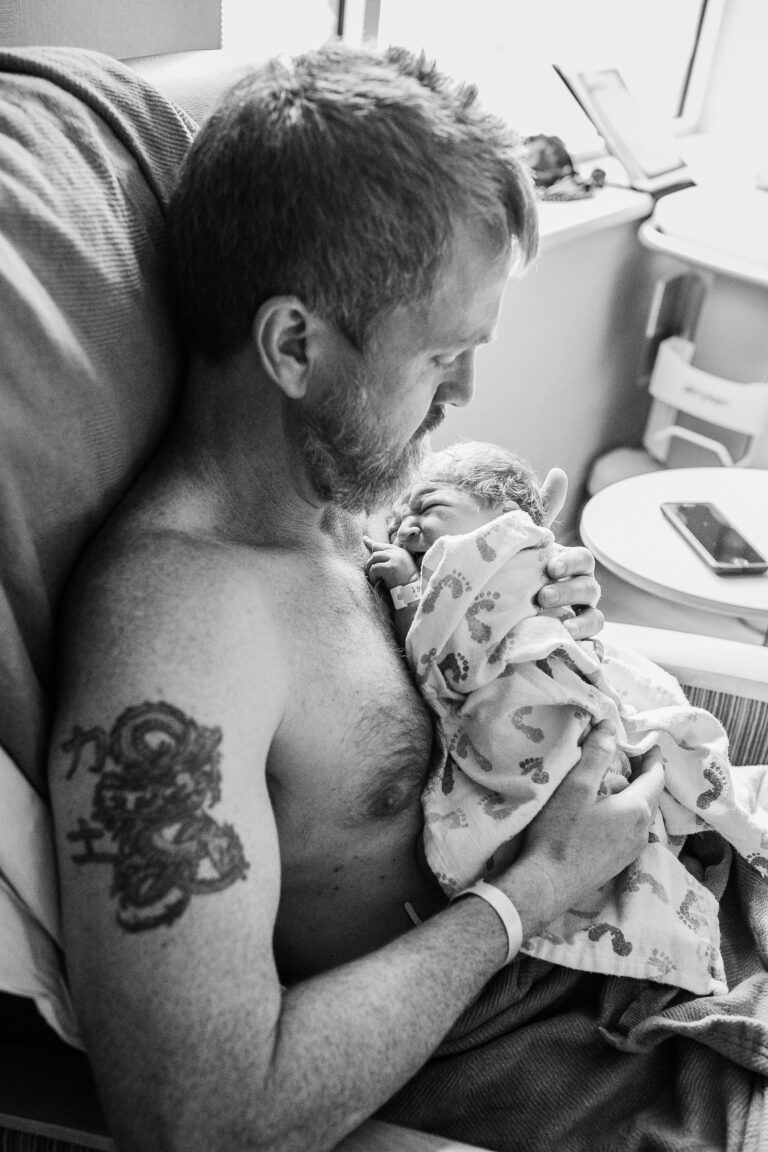 A new father gazes down protectively as he hold is crying newborn to his bear chest.