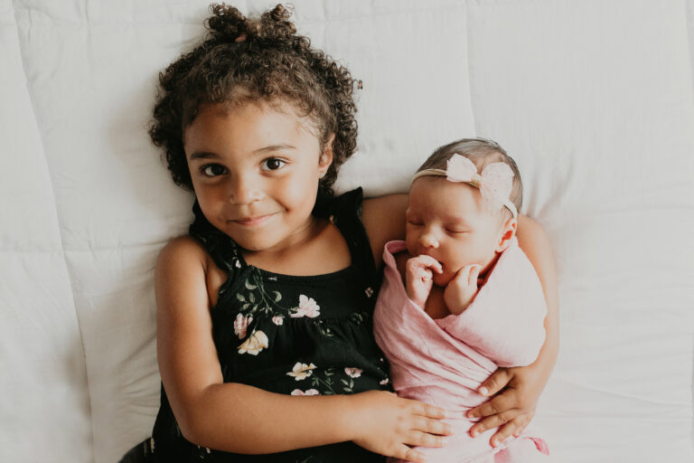 A young girl in tight curls casts a bright smile up at the camera as she proudly holds her new little sister.