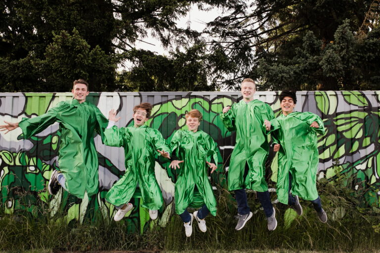 Five energetic seniors in emerald graduation gowns pose mid-air in before a graffitied train car.