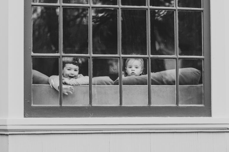 Two toddlers peak over couch cushions to peer out the living room window as a photographer captures their image in black and white.