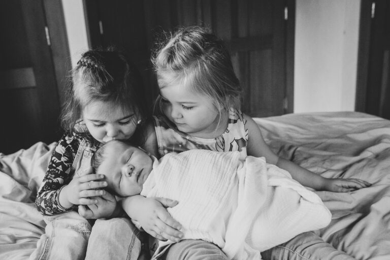 A four and a three year old gently cradle their newborn sibling as one sister plants a kiss on the babies head.