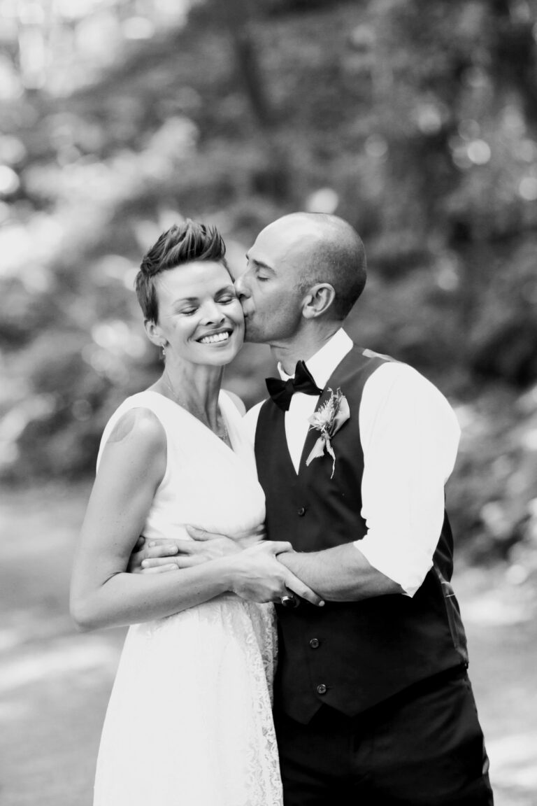 A bride beams as her groom pulls her into an embrace and plants a kiss on her cheek.