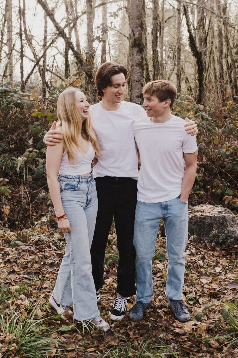 A trio of siblings in jeans and white tops stand arm in arm and share a laugh against a forest backdrop.