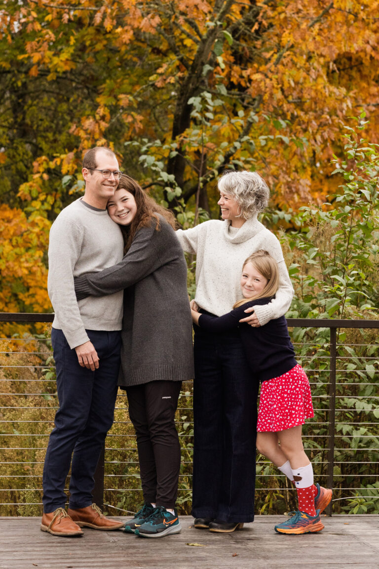 Two young daughters each embrace a parent on a balcony overlooking a forest of autumn leaves.