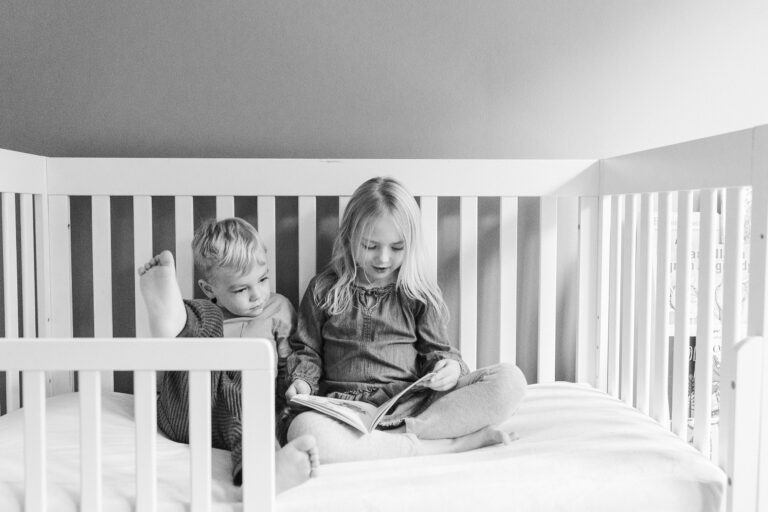 Two young children sit in a white toddler bed as the sister reads a book to her younger brother.