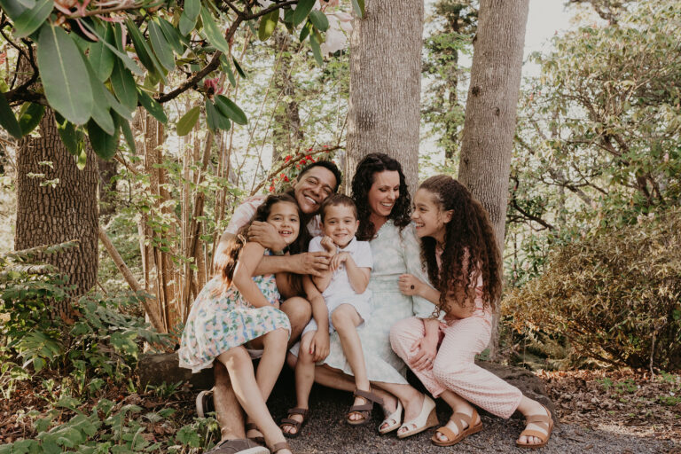 A beautiful young family, dressed in soft pastels, gathers together on a fallen tree in the forest, backlit my soft sun filtered through the trees.