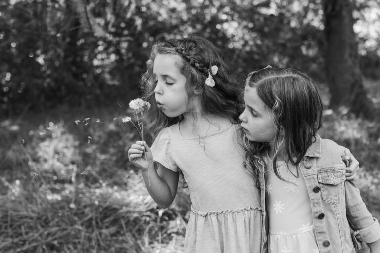 A young girl wraps an arm around her younger sister and demonstrates how to blow on a dandelion puff.