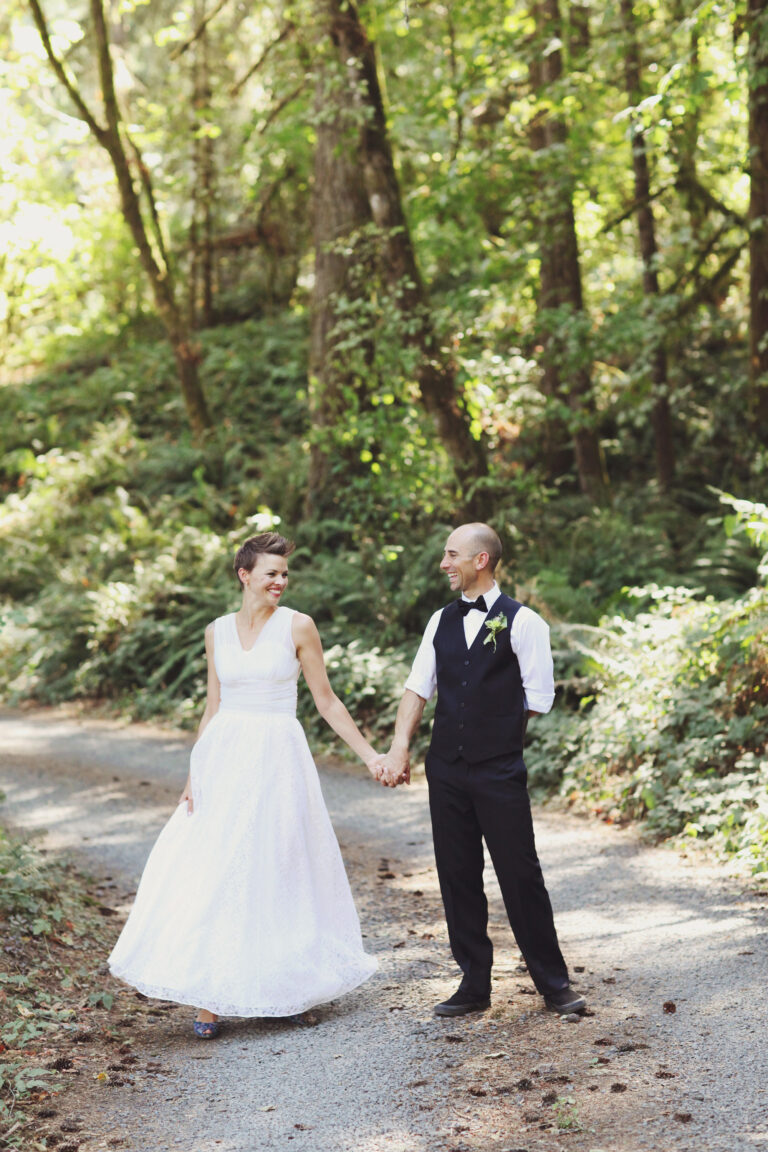 A bride twirls in her wedding gown along a forest trail as she holds hands with her smiling groom.