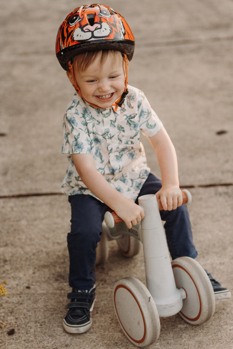 A delighted toddler wearing a tiger helmet scoots around a patio on his roller toy.