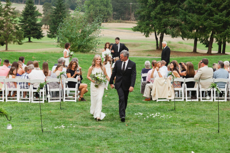 A newlywed couple stroll clasp hands and stroll down the isle at the end of their ceremony as the wedding attendees look on.