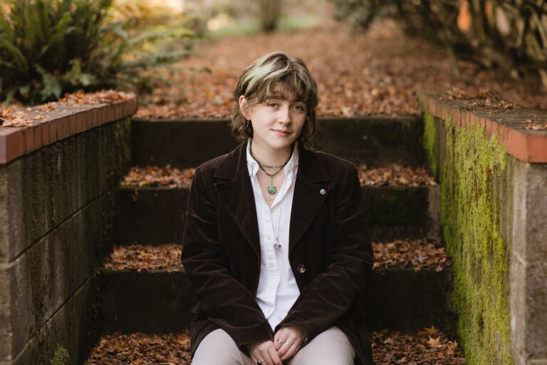 A high school senior girl sits on steps surrounded by colorful autumn leaves, enjoying the crisp fall day.