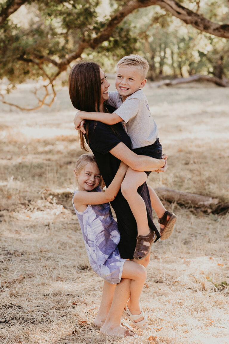 Woman hugging two smiling children in a park.