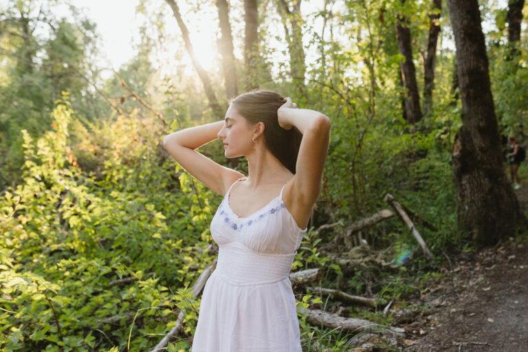 A woman in a flowing white dress stands gracefully among the trees in a serene forest setting.