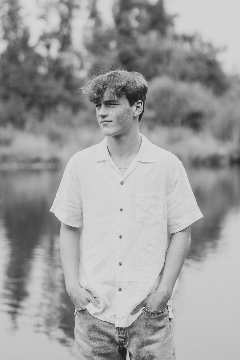 A young man wearing a white shirt stands beside a tranquil lake, taking in the calm atmosphere and scenic view.