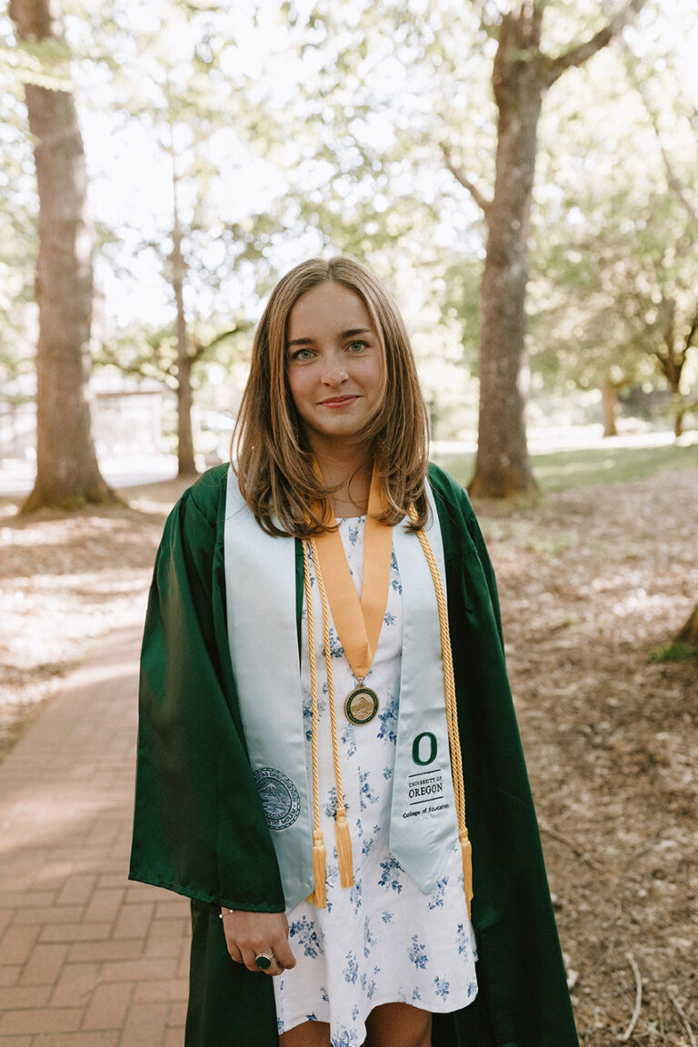 A proud girl in a graduation gown and medal smiles in a sunny park, celebrating her achievement.