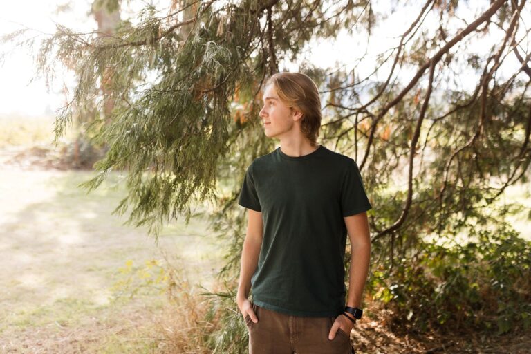 A young man stands confidently in front of a large tree, enjoying the serene outdoor setting.