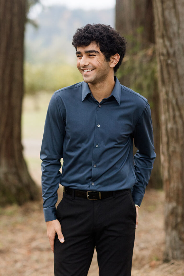A young man dressed in a blue shirt and black pants smiles, exuding a relaxed and fashionable vibe.