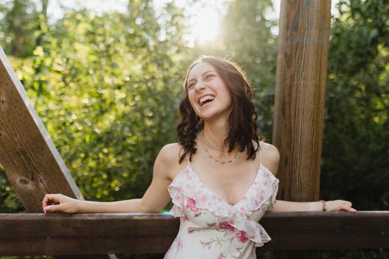 A woman in a floral dress beams with joy while standing on a charming wooden bridge surrounded by nature.